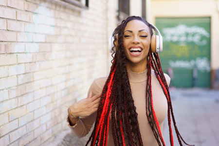 A vibrant young woman with long, braided hair and stylish headphones radiates joy and happiness as she smiles in a lively urban setting, embodying the spirit of music and happinessの写真素材