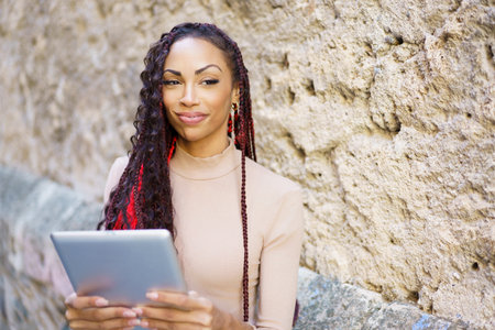 A stylish woman with long, braided hair relaxes against a textured stone wall, engaging with a tablet, blending modern technology with her confidence and embracing contemporary leisureの写真素材