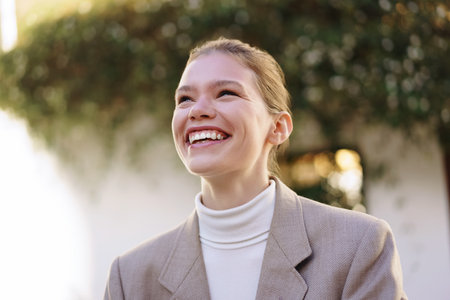 A cheerful young woman smiles brightly in a relaxed outdoor setting, embodying happiness,...