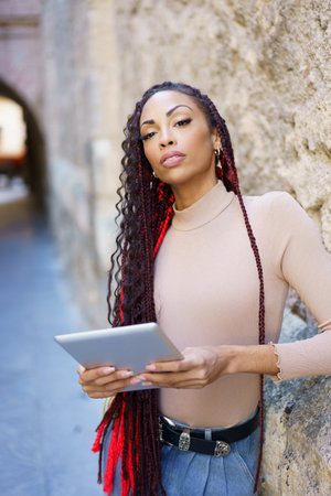 A poised and stylish woman confidently stands against a textured wall, holding a tablet, exuding an air of poise and sophistication in a bustling urban environment filled with life and creativityの写真素材