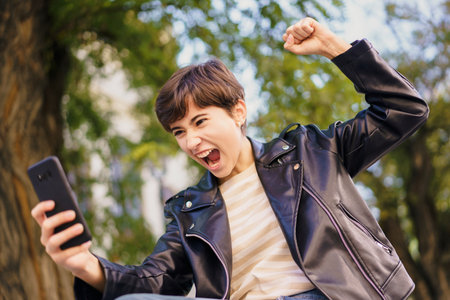 In a vibrant park, a young person joyfully celebrates their success by sharing their enthusiasm with friends via smartphone, highlighting the significant impact of modern technology on our livesの写真素材