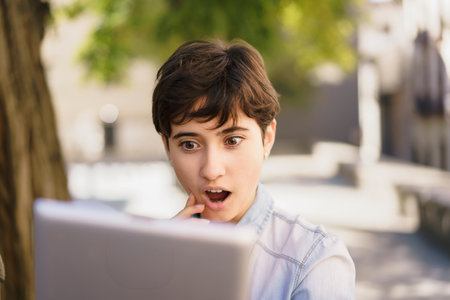 A young woman demonstrates a look of surprise and wonder while using her laptop outdoors, highlighting a captivating moment filled with curiosity, engagement, and exploration of technologyの写真素材
