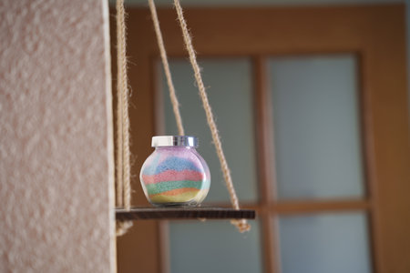 hanging glass jar filled with sand, layered pastel sand resting on tiny wooden swing suspended by...