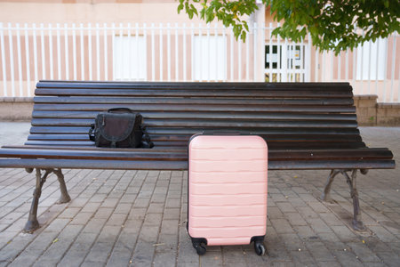 Pink suitcase and backpack on bench awaiting traveler pickup empty city street, leafy shade, soft morning light, cobblestone pavement, subtle urban calm,の写真素材