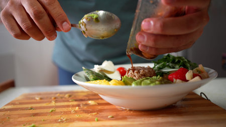 Closeup hands drizzling dressing over salad bowl on wooden cutting board, cherry tomatoes and cucumber slices mingling with mixed greens, spoon glintingの写真素材