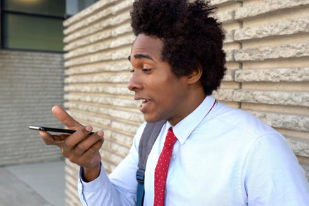 Black entrepreneur sending audio message outdoors, white shirt with red tie, smartphone held close...