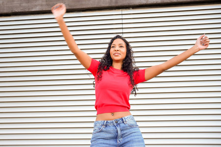 Young woman with arms raised joyful expression in front of metal shutter wearing red top and jeans,...