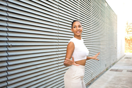 Smiling woman jogging beside metal wall, sunlit urban corridor wearing white crop top and beige leggings midstride with confident smile holding phone armbandの写真素材