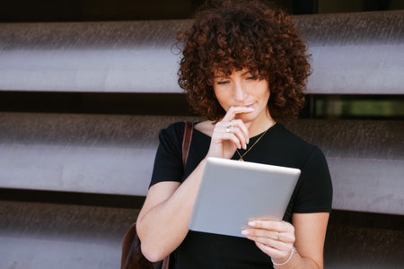 Woman pondering over tablet screen outdoors, finger near lips, thoughtful expression, reviewing...