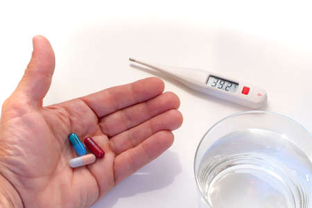 digital thermometer showing fever values, hand holding tree pills and water glassの写真素材