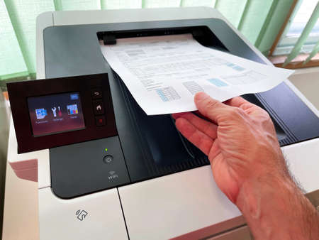 hand holding a sheet of paper with a freshly printed report on a modern wifi multifunction printer with a touch screen control in an office, horizontalの写真素材