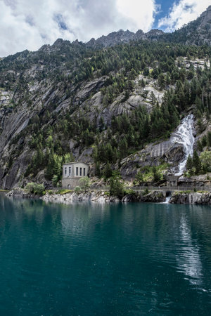 Cavallers reservoir between high mountains, a waterfall and a building of the hydroelectric power station, river Noguera de Tor in Ribagorza, Boi Taull valley, Lleida Pyrenees, Spain, verticalの写真素材