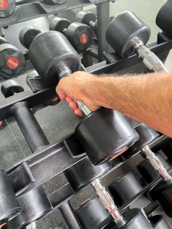 a hand of a man holding a black dumbbell from a rack in a gymnasium during an indoor training session, verticalの写真素材
