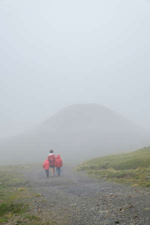 a young mother with her two children holding hands strolls through a mountain landscape on a foggy day, copy space, verticalの写真素材