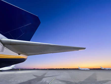 tail of an aircraft, on the ground of an airport, with two airplanes in the background and a sunset sky in the backgroundの写真素材