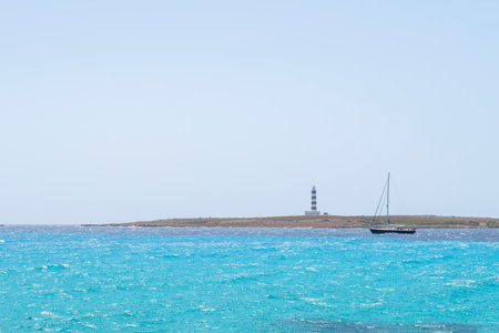a sailboat skirts a cape with a blue and white lighthouse, on a sea of turquoise waters, clear sky, Isla del Aire, Punta Prima, Menorca, Spain, copy spaceの写真素材