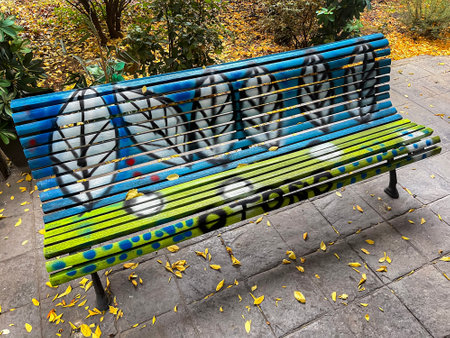 a park bench painted in yellow and blue with a leaf and a message in Spanish with the meaning of autumn, fallen leaves from the trees all around it, colorful bench, horizontalの写真素材