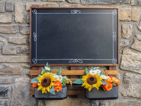 an empty black blackboard hanging on a stone wall with some decorative flowers below it, copy space, empty menu board, horizontalの写真素材