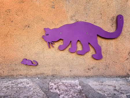a concrete orange wall with purple wooden cat stalking a small mouse , funny purple cat chasing a scaredy small mouse in a cement wall, street decoration, horizontalの写真素材