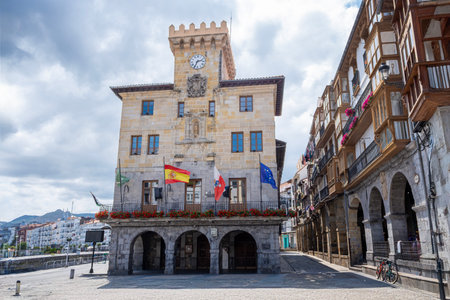 main facade of the town hall of Castrourdiales, with the flags waving in the wind, with its empty square of people, and its typical arcaded arcades, on a cloudy summer dayの写真素材