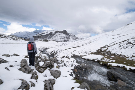 Hiker with a red backpack stands on snowy rocks, overlooking a mountain stream and snow-covered peaks and a mountain river under a dramatic sky in the Pyreneesの写真素材
