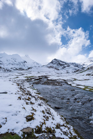 Snowy mountain landscape with a clear stream flowing through a valley under a partly cloudy skyの写真素材
