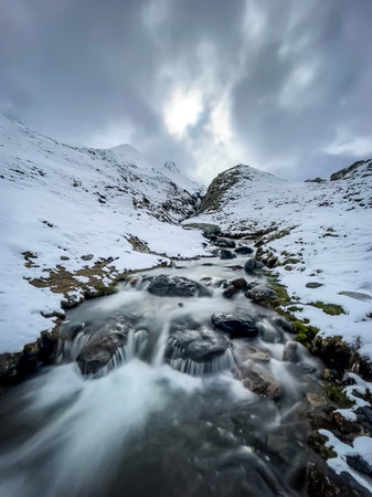 a torrent of mountain snowmelt flows between the slopes of a small valley on top of a snow-capped...