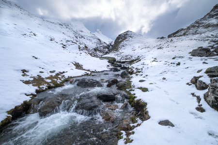 Snow-covered mountains with a clear stream flowing through rocky terrain under a cloudy sky,...
