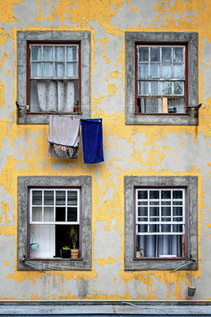Old facade in Porto with peeling yellow paint, four rustic windows, hanging laundry, and small potted plants creating an authentic urban atmosphereの写真素材