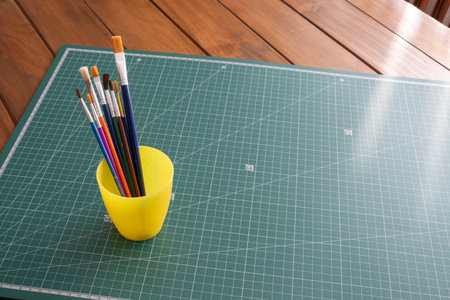 A collection of multicolored paint brushes in a yellow cup sits on a green cutting mat next to a...