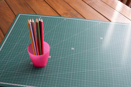 A collection of multicolored pencils in a pink cup sits on a green cutting mat next to a wooden table. Copy space on the mat is ideal for art design or creative concepts.の写真素材