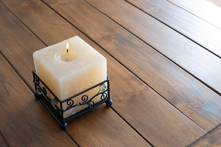 A lit square candle in an elegant metal holder on a wooden table, creating a warm and peaceful atmosphere ideal for relaxation and calm momentsの写真素材