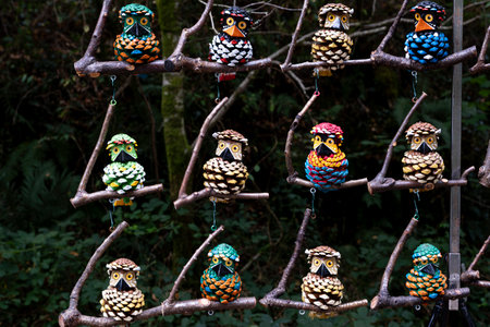 Colorful handmade funny owl figures crafted from pine cones hang on wooden branches in a forest setting, creating a vibrant and playful outdoor displayの写真素材
