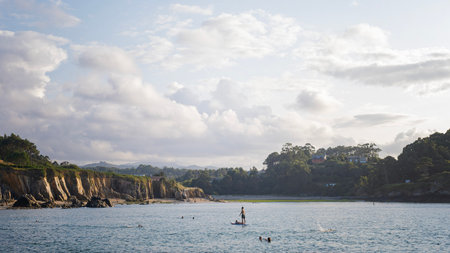 Asturias, Spain; August 20 2025: Coastal scene from Porcia beach in Asturias with swimmers and a person on a paddleboard in calm waters, framed by cliffs, trees, and soft evening light under a skyのeditorial素材