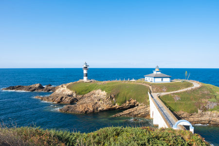Scenic landscape of Isla Pancha in Ribadeo, Spain, featuring two historic lighthouses on a rocky island connected by a bridge under a clear blue skyの写真素材