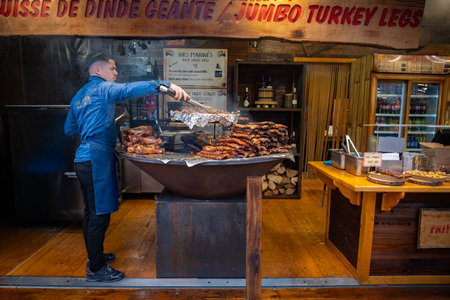 Paris, France; December 28 2025: A chef grills large quantities of ribs and turkey legs at an outdoor Christmas market stall in Paris. Traditional festive street food prepared on a large circularのeditorial素材