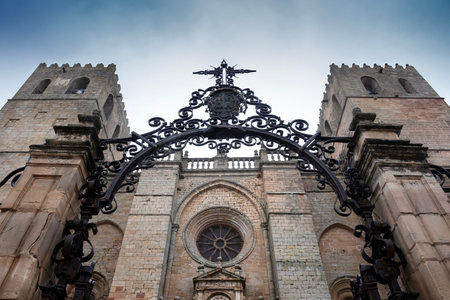 Low angle view of the Romanesque facade and towers of Siguenza Cathedral through an ornate wrought iron arch. Historic medieval stone architecture in Guadalajara, Spain, under a dramatic skyの写真素材