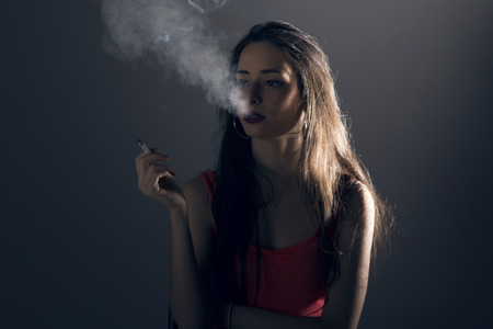 Beautiful young woman with a cigarrette, exhaling smoke, over grey background.の写真素材