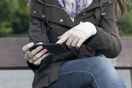 Woman with a injured wrist wrapped in an bandage, using a smartphone, outdoors.の写真素材