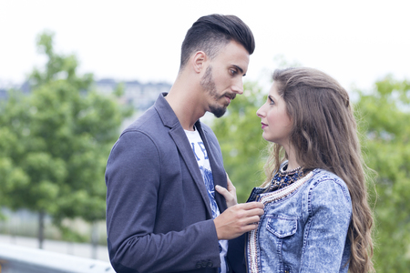 Young couple in love outdoors, in the city near a group of trees. They are holding their jackets and looking at each other.の写真素材