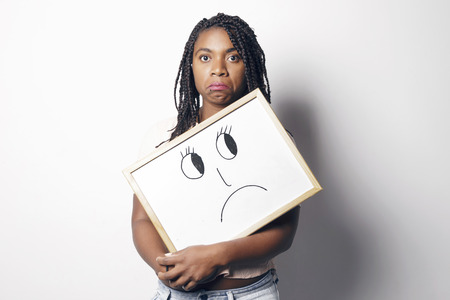Sad young african woman holding an unhappy smiley white board, over a white background.の写真素材