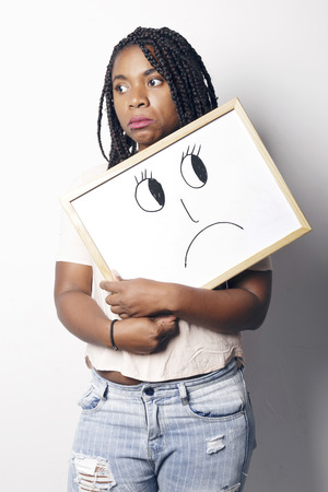 Sad young african woman holding an unhappy smiley white board, over a white background.の写真素材