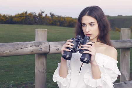 Young brunette woman watching with binoculars, looking and exploring her future. Outdoors. Shallow depth of field.の写真素材