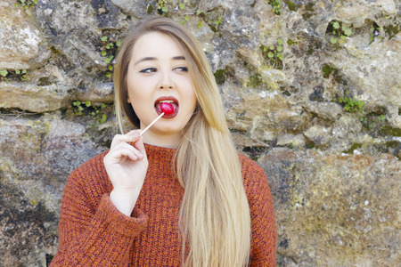 Cheerful young girl on orange sweater, with candy on stick, Chupa Chups, outdoors on a sunny day...