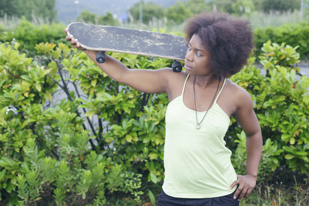 African woman with a skateboard, posing in a park, over a green bush, outdoors. Slightly depth of field. Focus on her eyes.の写真素材
