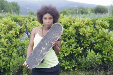 African woman with a skateboard, posing in a park, over a green bush, outdoors. Slightly depth of field. Focus on her eyes.の写真素材