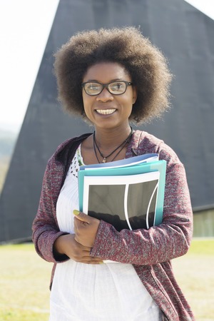 African clever girl standing on the street, on a sunny day, holding a few folders and books.の写真素材