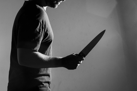 Man in black t-shirt, standing holding a knife. Indoors. Converted to black and white, grain added.の写真素材