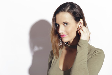 Young woman with a wireless earphone,  connectivity with her smartphone. Indoors, over a white...