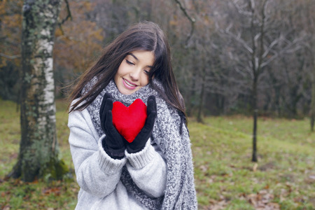 Young brunette woman holding a heart shaped cushion, outdoors, in winter mountains. Valentines day concept. Love.の写真素材